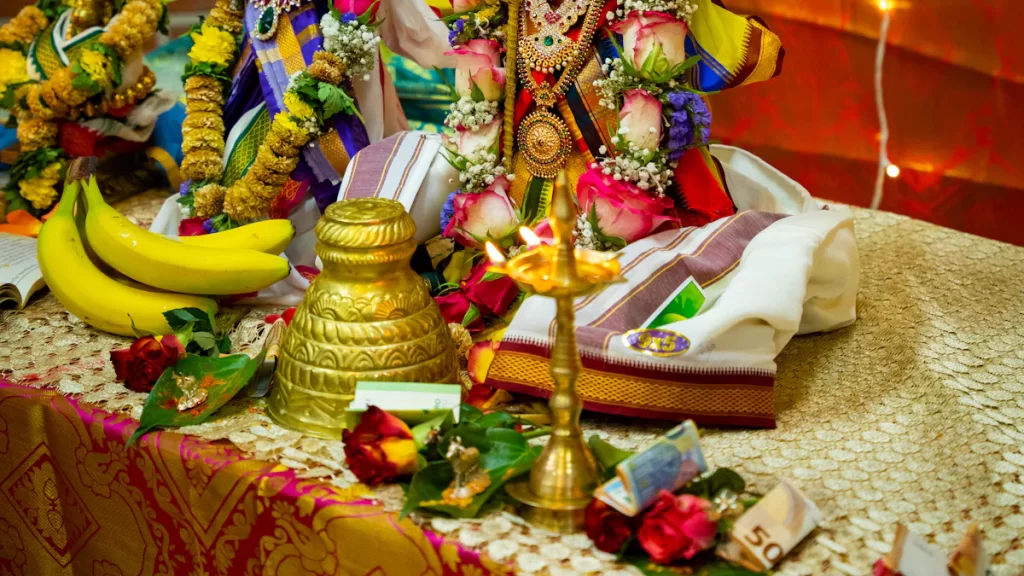 A traditional altar display with a brass lamp, bananas, floral garlands, a decorated deity, currency notes, and red roses on a patterned cloth